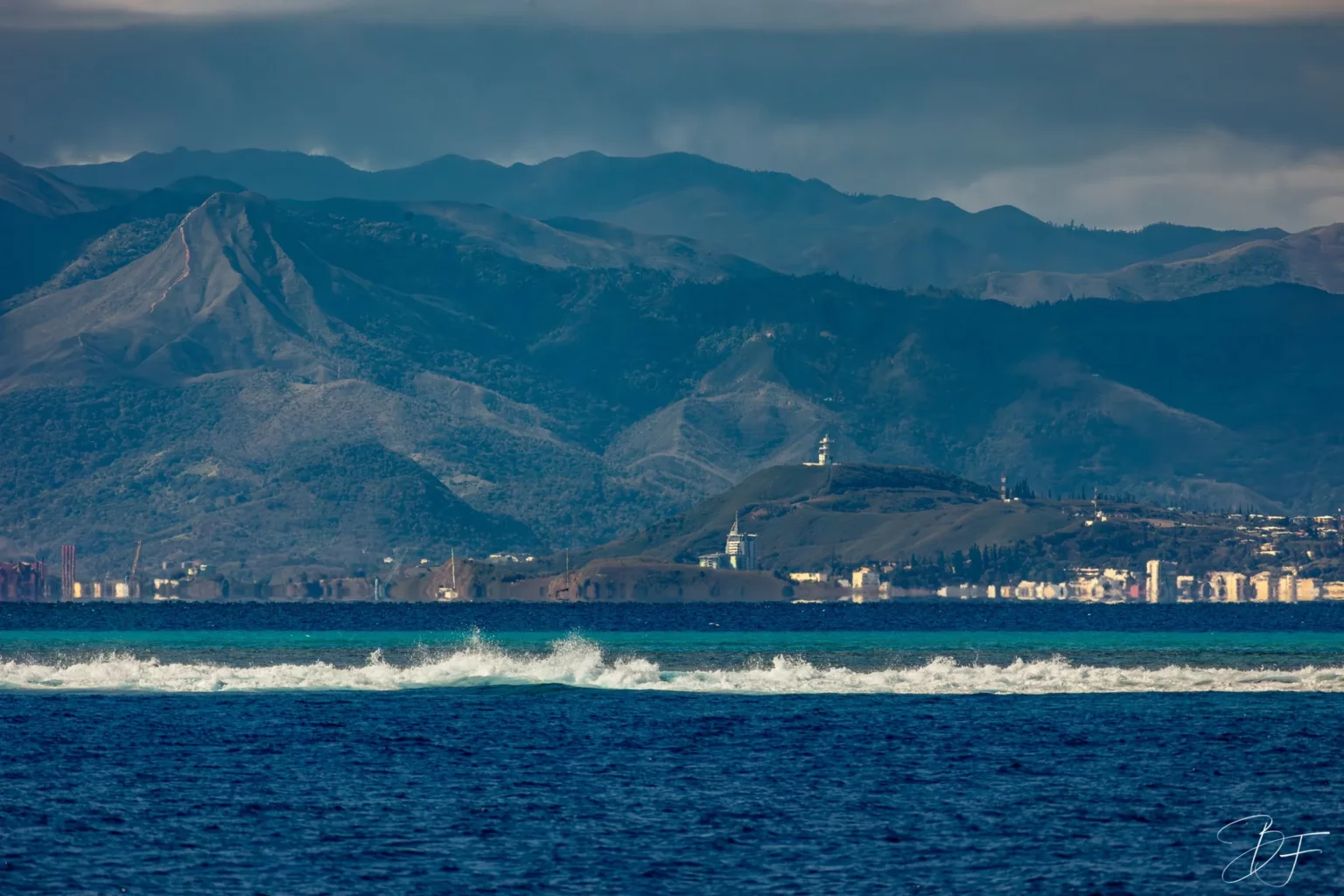 La côte de Nouméa et ses montagnes vues depuis le lagon, une vague déferlant sur le récif